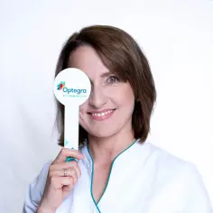 Uśmiechnięta kobieta w białym fartuchu zakrywa jedno oko tabliczką z logo "Optegra Eye Health Care", doświadczając chwilowej utraty ostrości widzenia podczas badania.