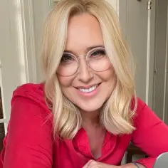 Uśmiechnięta blondynka w okularach, które odmładzają. Czerwona koszula, jasne wnętrze.