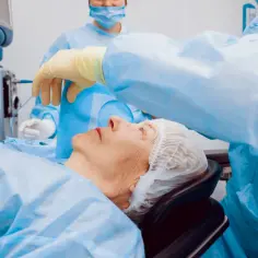 Pacjentka leży na stole operacyjnym, przygotowana do zabiegu. Lekarz w rękawiczce delikatnie dotyka jej twarzy. To moment przed operacją zaćmy, po której pacjentka otrzyma zwolnienie lekarskie L4.