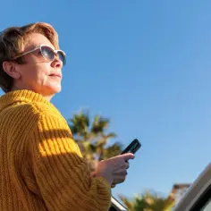 Kobieta w żółtym swetrze i okularach przeciwsłonecznych trzyma telefon. Jaskra słońca odbija się w jej okularach, a palmy w tle sugerują ciepły dzień.