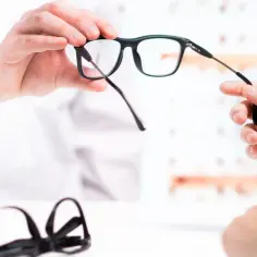 Dwie ręce przekazują sobie czarne okulary. Czy okulary są refundowane? W tle widać inne oprawki.