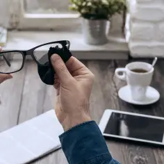 Mężczyzna czyści okulary z antyrefleksem czarną ściereczką. Na biurku leży laptop, tablet i filiżanka kawy.