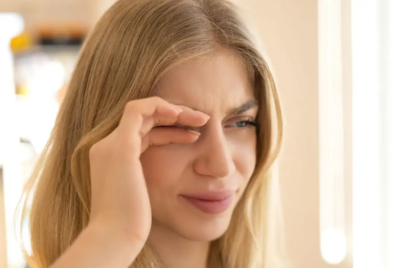 Kobieta z blond włosami pociera oko, wyraźnie odczuwając problem ze skupieniem wzroku.