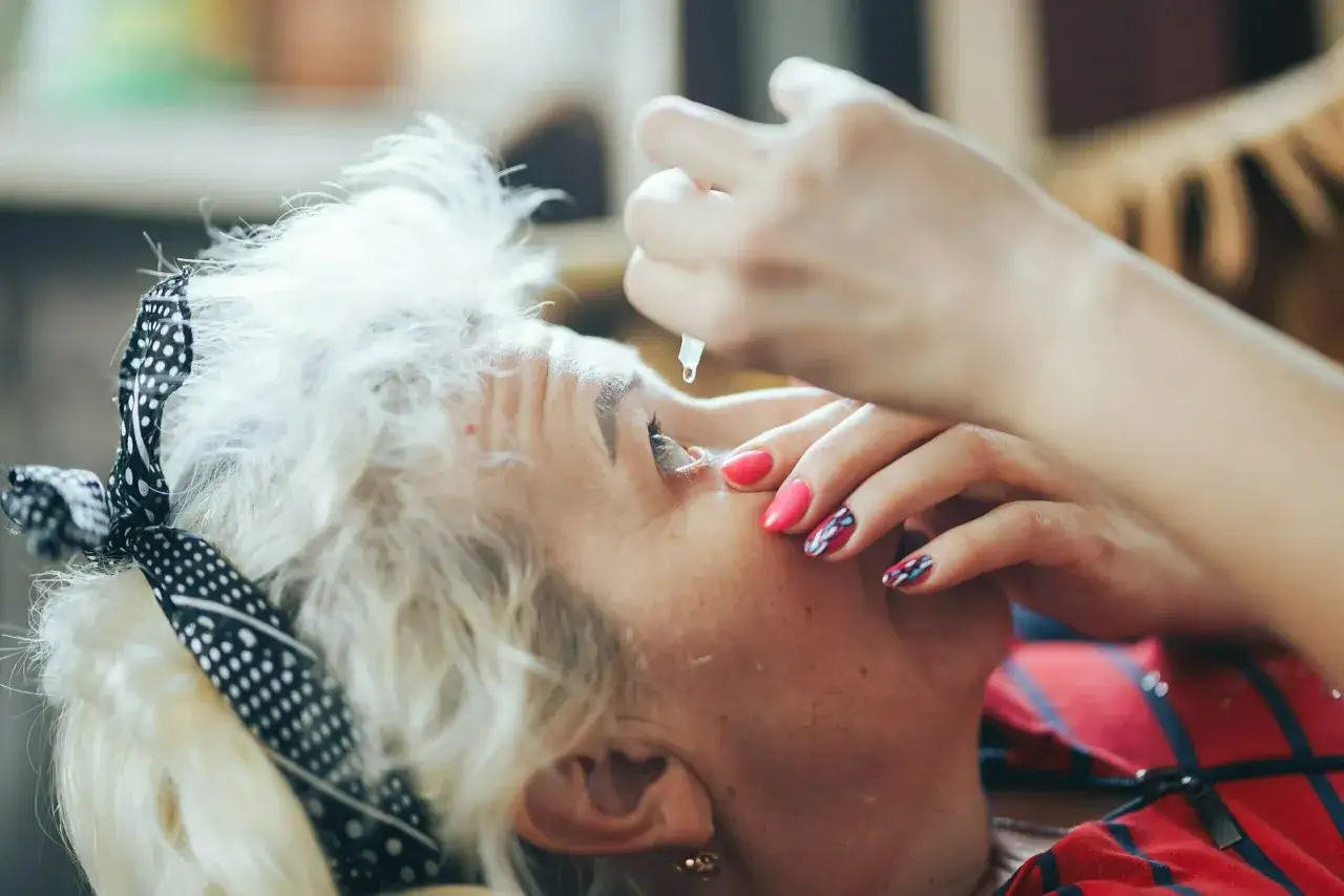 Krople przed operacją zaćmy podawane starszej kobiecie. Dłoń z kolorowym manicure aplikuje lek.